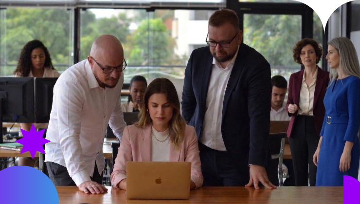 Três pessoas olhando um computador sobre a mesa, sendo dois homens nas pontas e uma mulher no centro, eles estão em uma sala de reunião e com pessoas ao seu redor na sala ao fundo